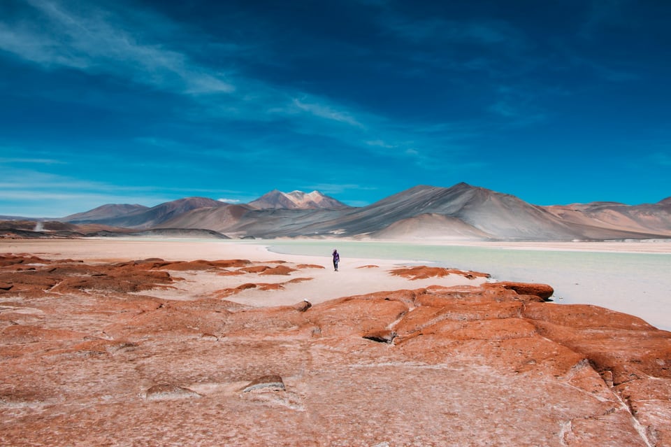 Red mineral ground, a pale lagoon and volcanic peaks beneath a deep blue sky in the Atacama Desert