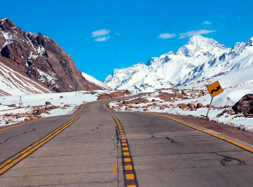 Paved mountain road curving between red rock and snow-covered peaks at Paso Los Libertadores
